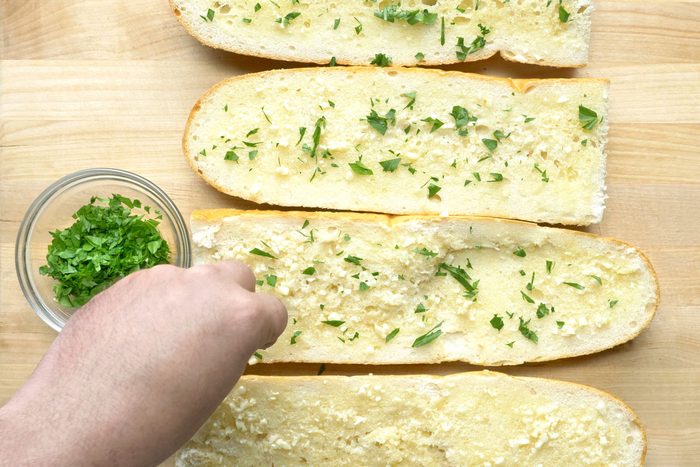 overhead shot of four slices of bread topped with garlic butter and parsley; the bread slices are arranged in a row on a wooden cutting board; a hand is holding a small glass bowl filled with chopped parsley; the hand is reaching down to sprinkle parsley on top of the bread slices