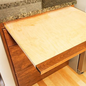 Maple wood cutting board pulls out from between a marble kitchen counter and the wooden cabinets below