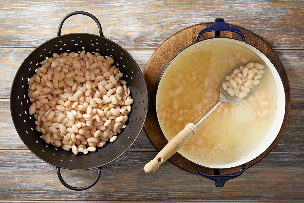A colander filled with white beans sits next to a pot containing beans in broth. Both are placed on a round wooden board on a rustic wooden table. A slotted spoon rests on the pot.