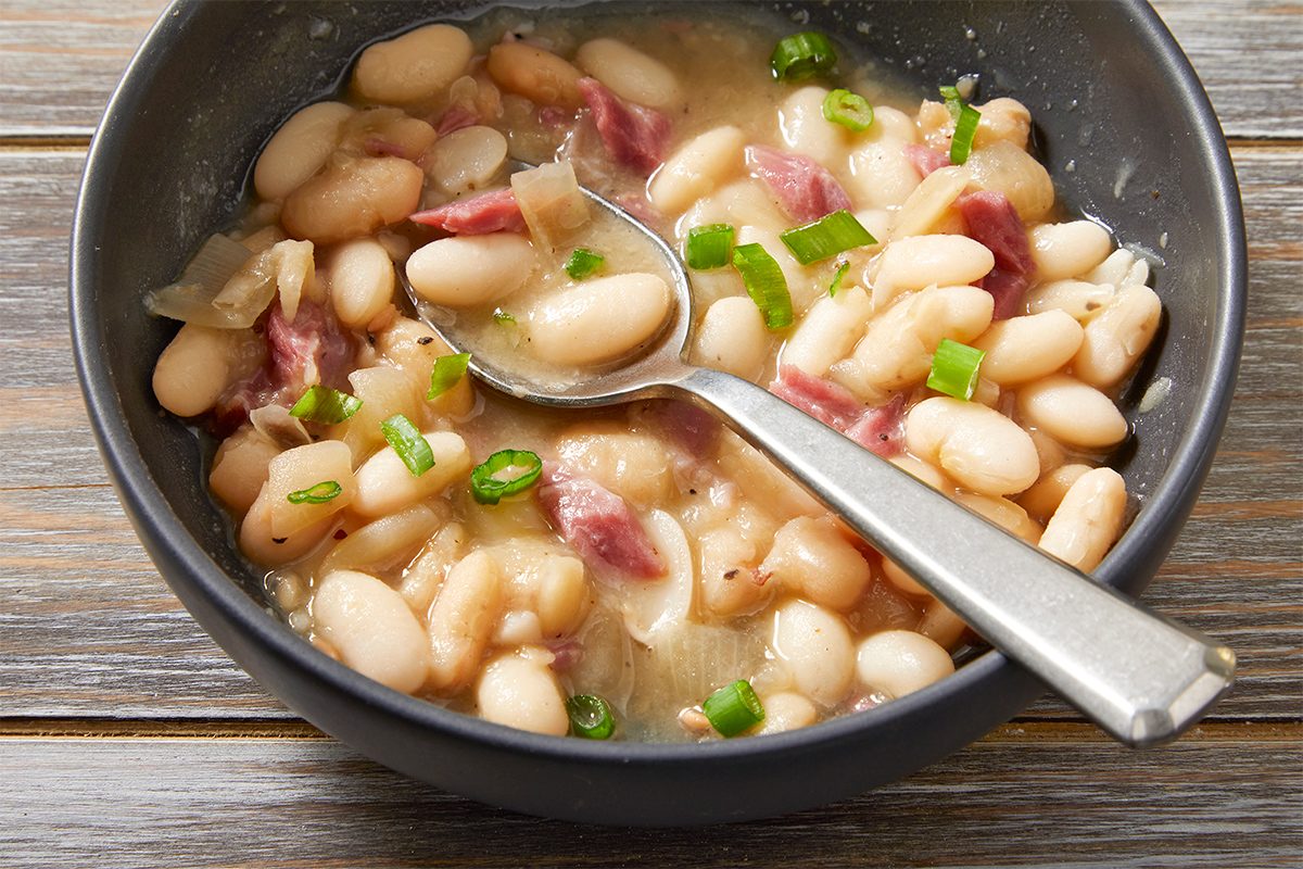 A bowl of white bean soup with chunks of ham and sliced green onions. A spoon rests inside the bowl. The soup is served in a dark gray bowl on a wooden surface.