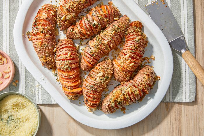 A white platter holds several roasted Hasselback sweet potatoes topped with a breadcrumb and herb mixture. A small bowl of grated cheese and a knife are nearby on a striped cloth.