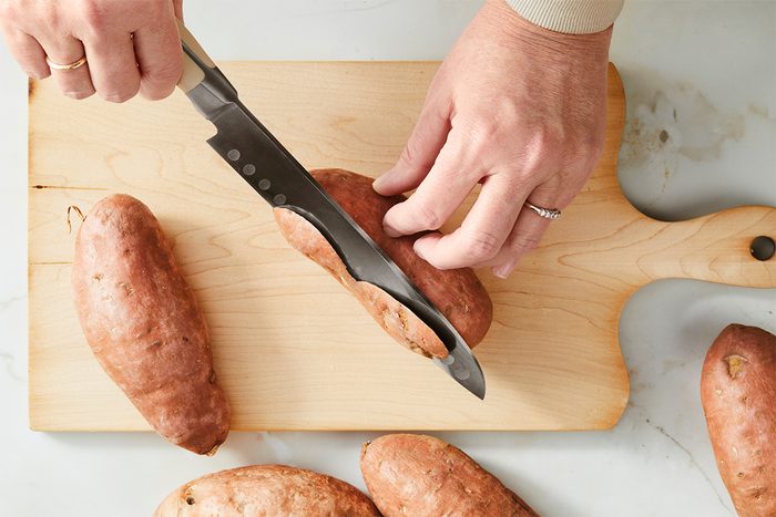 A person cutting sweet potatoes on a wooden cutting board with a large knife. The board is on a light-colored countertop, and several uncut sweet potatoes are placed around the cutting board.