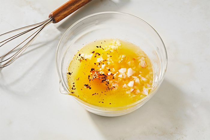 A clear glass bowl contains a yellow mixture of oil, finely chopped garlic, and black pepper, set on a light-colored marble surface. A whisk with a wooden handle lies beside the bowl.