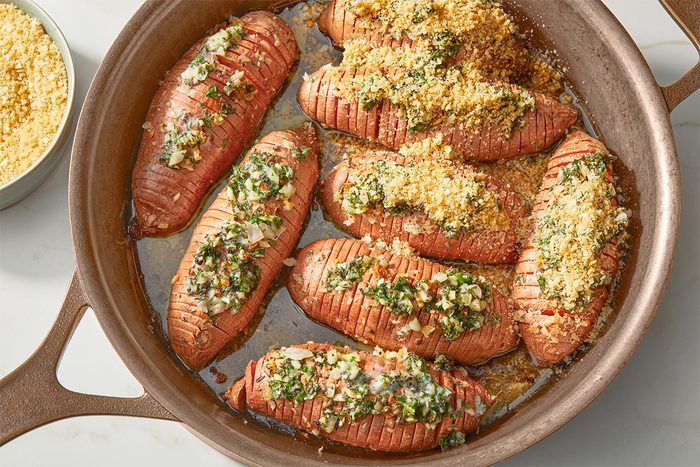 Baked sweet potatoes in a roasting pan, sliced in a hasselback style and topped with a mixture of herbs, breadcrumbs, and seasoning. A small bowl of breadcrumbs is visible in the background.