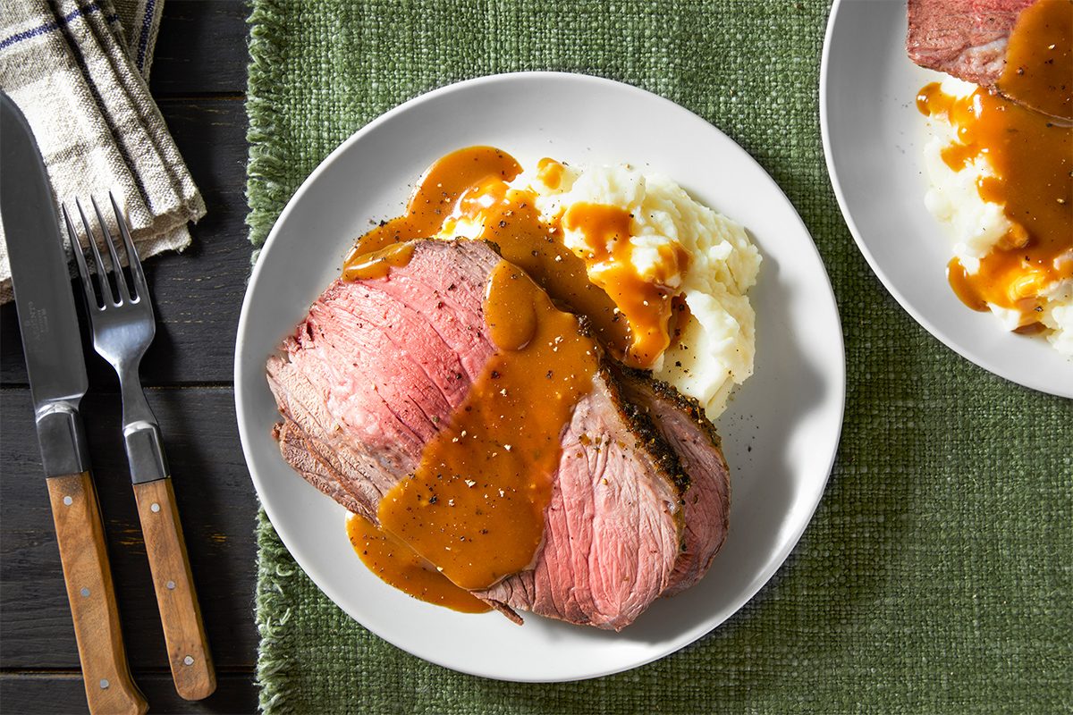 Top view shot of Herb-Crusted Roast Beef; served on plates with mashed potatoes; serve with gravy; knife and fork; napkin; table cloth; dark wooden surface;