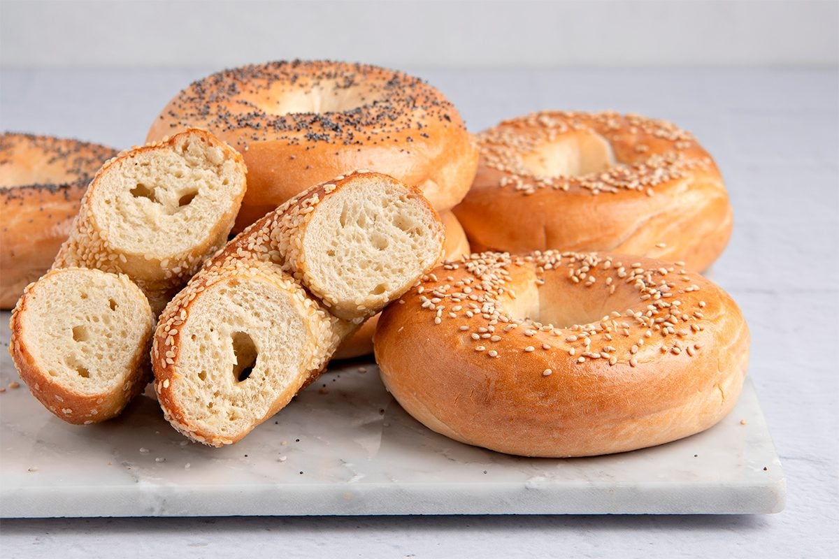 A variety of bagels are displayed on a white marble surface. Some are topped with sesame seeds, others with poppy seeds, and one is sliced in half to show the texture. The background is a light gray.