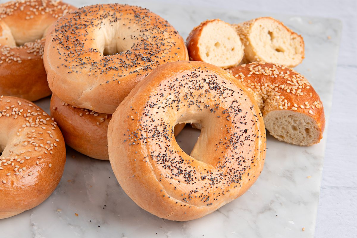 A variety of bagels are arranged on a marble surface. They include plain, poppy seed, and sesame seed toppings. A few bagels are sliced in half, showcasing their soft interior.