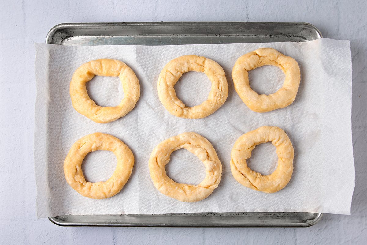 Six unbaked bagels arranged on a baking sheet lined with parchment paper, placed on a light-colored surface.