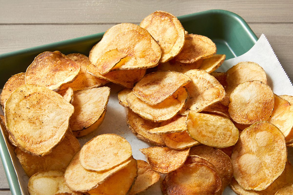 A green tray filled with crispy, golden-brown potato chips. The chips are slightly overlapping, showing their irregular, round shapes. A paper towel is partly visible under the chips, absorbing some oil. The background is a wooden surface.