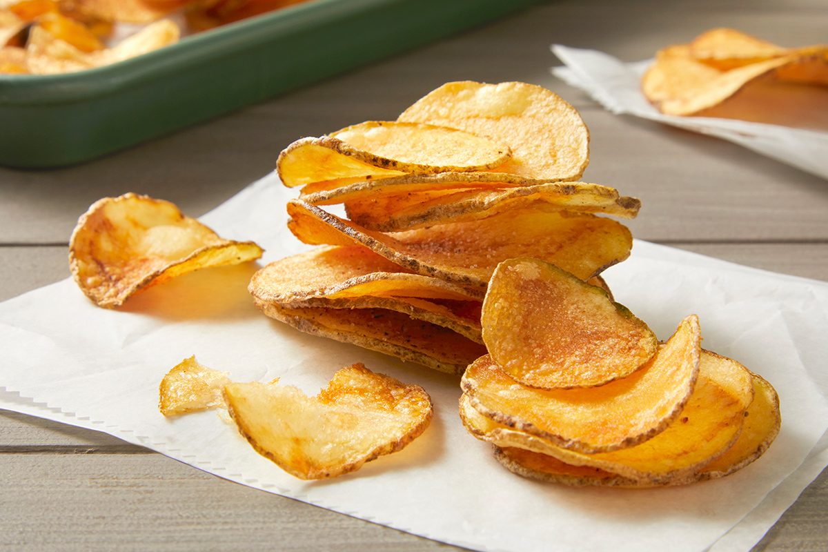 A stack of crispy potato chips sits on white parchment paper on a wooden surface. Some chips are scattered nearby, with a green tray filled with more chips visible in the background.