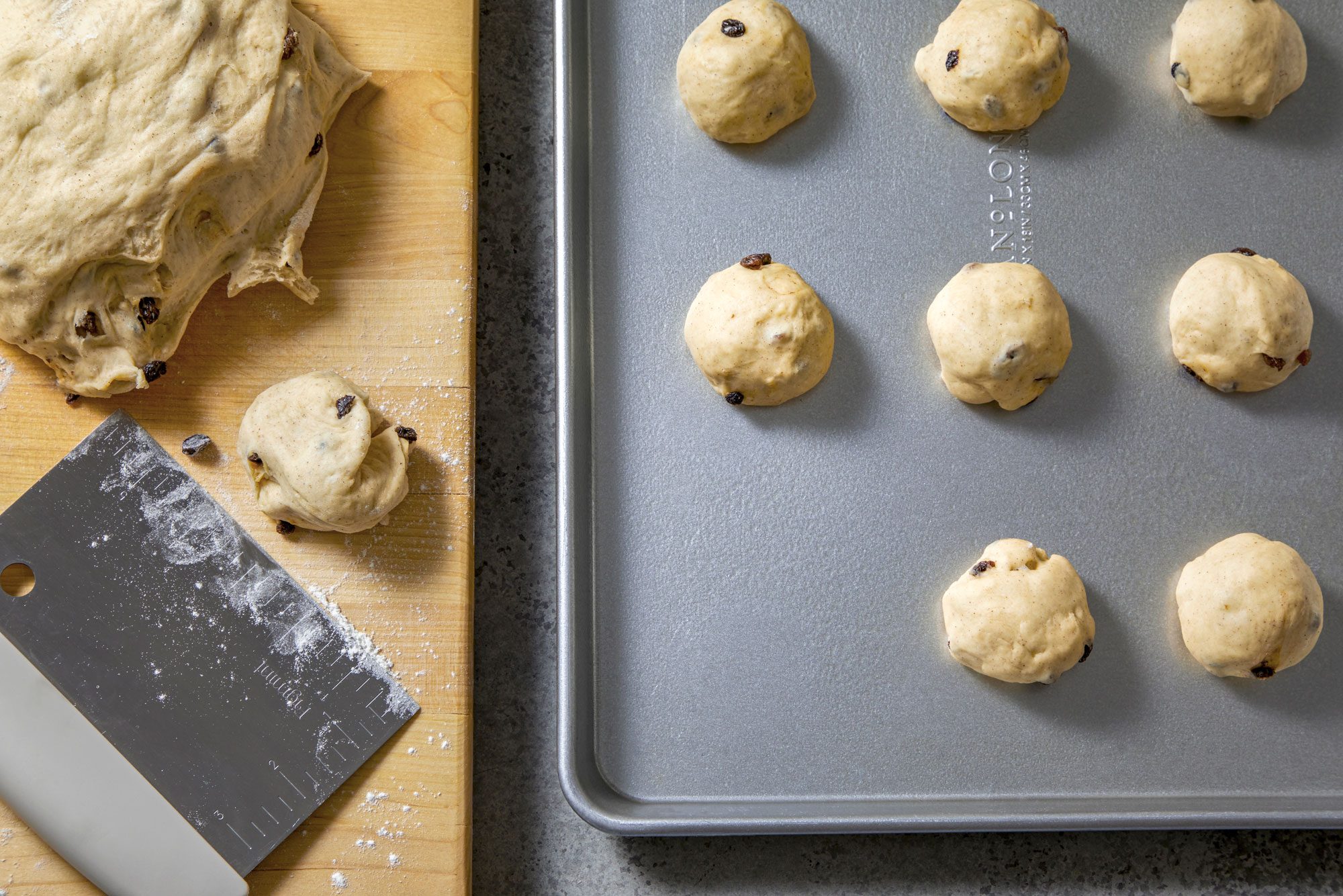 overhead shot of a baking preparation process; on the left side of the image there is a wooden cutting board with a large piece of dough with raisins on it; there is also a smaller ball of dough with raisins on it and a metal scraper with a white handle; there is a metal baking sheet on the right side of the image; there are seven balls of dough with raisins on the baking sheet