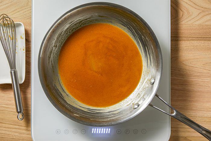 A stainless steel pan on an induction cooktop contains a vibrant orange sauce. A whisk rests beside the cooktop on a white dish, over a wood-grain countertop.