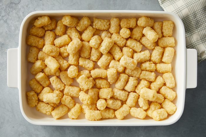 A white rectangular dish filled with evenly spread tater tots is placed on a gray surface. A portion of a plaid cloth is visible at the top right corner of the image.