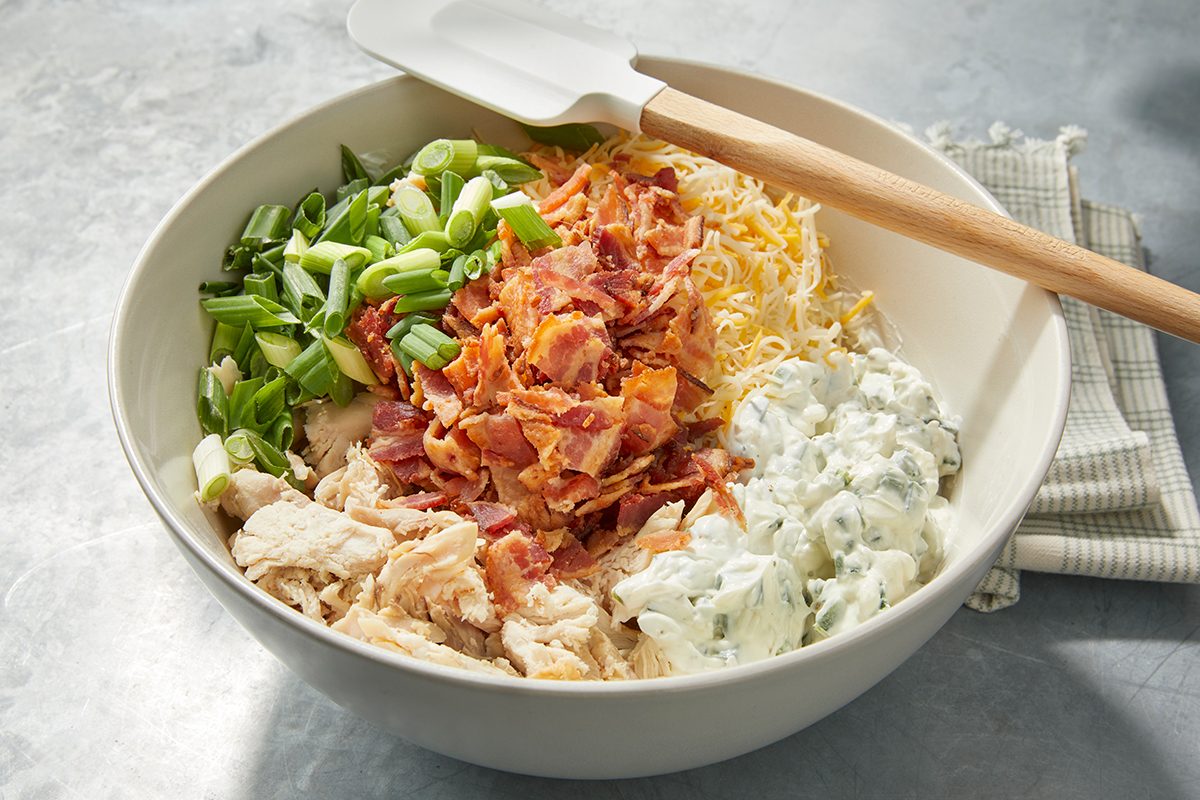 A large bowl contains chopped cooked chicken, crispy bacon bits, shredded cheese, chopped green onions, and a creamy sauce with herbs. A white spatula rests on the bowl's edge, and a cloth lies nearby on the table.