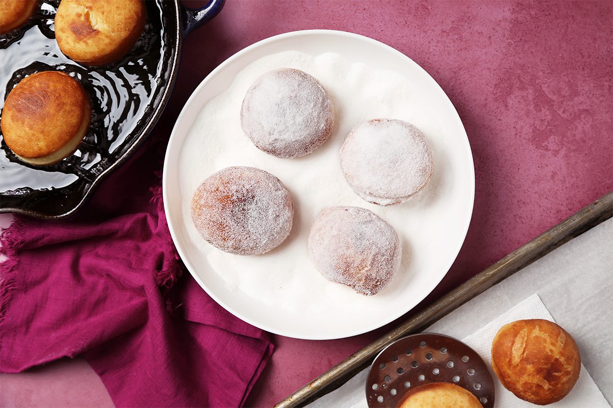 A white plate with four powdered beignets sits on a pink surface, accompanied by a maroon cloth. Nearby, a dark pan contains more golden beignets, and a small brown polka-dot bowl holds another pastry.