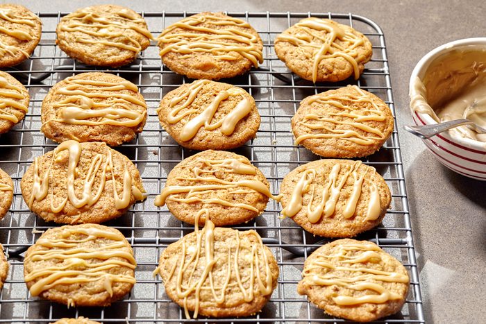 Horizontal AP Pull shot of a cooling rack with freshly baked cookies; the cookies are iced with a caramel glaze, and the glaze is still wet and shiny; a small bowl of caramel glaze is on the right side of the image, and a spoon is in the bowl