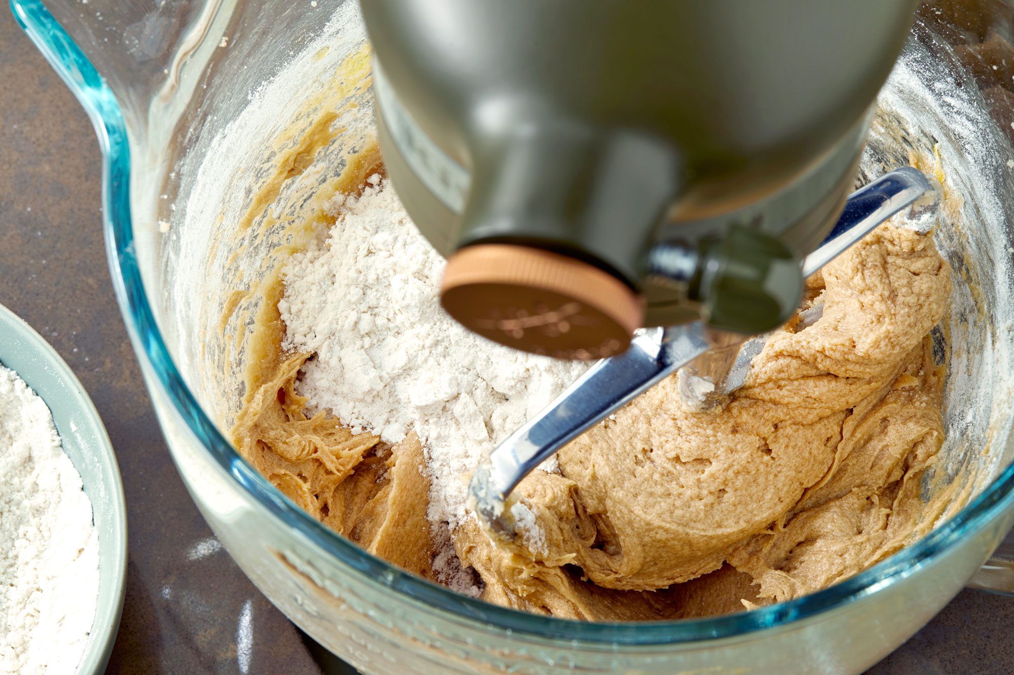 a close-up view of a kitchen mixer's bowl during the baking process; the bowl is filled with a mixture of batter and flour, while the mixer's metal paddle sits in the batter, partially submerged