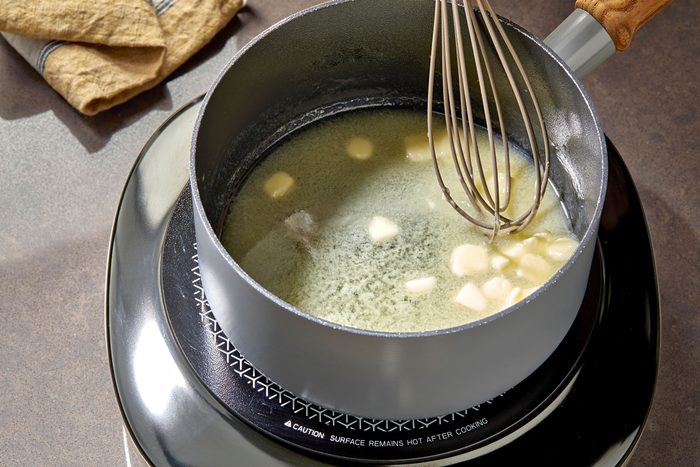 3/4th shot of a silver saucepan sitting on a black electric stovetop burner, the burner is on and the pan is filled with melted butter and a wooden-handled whisk