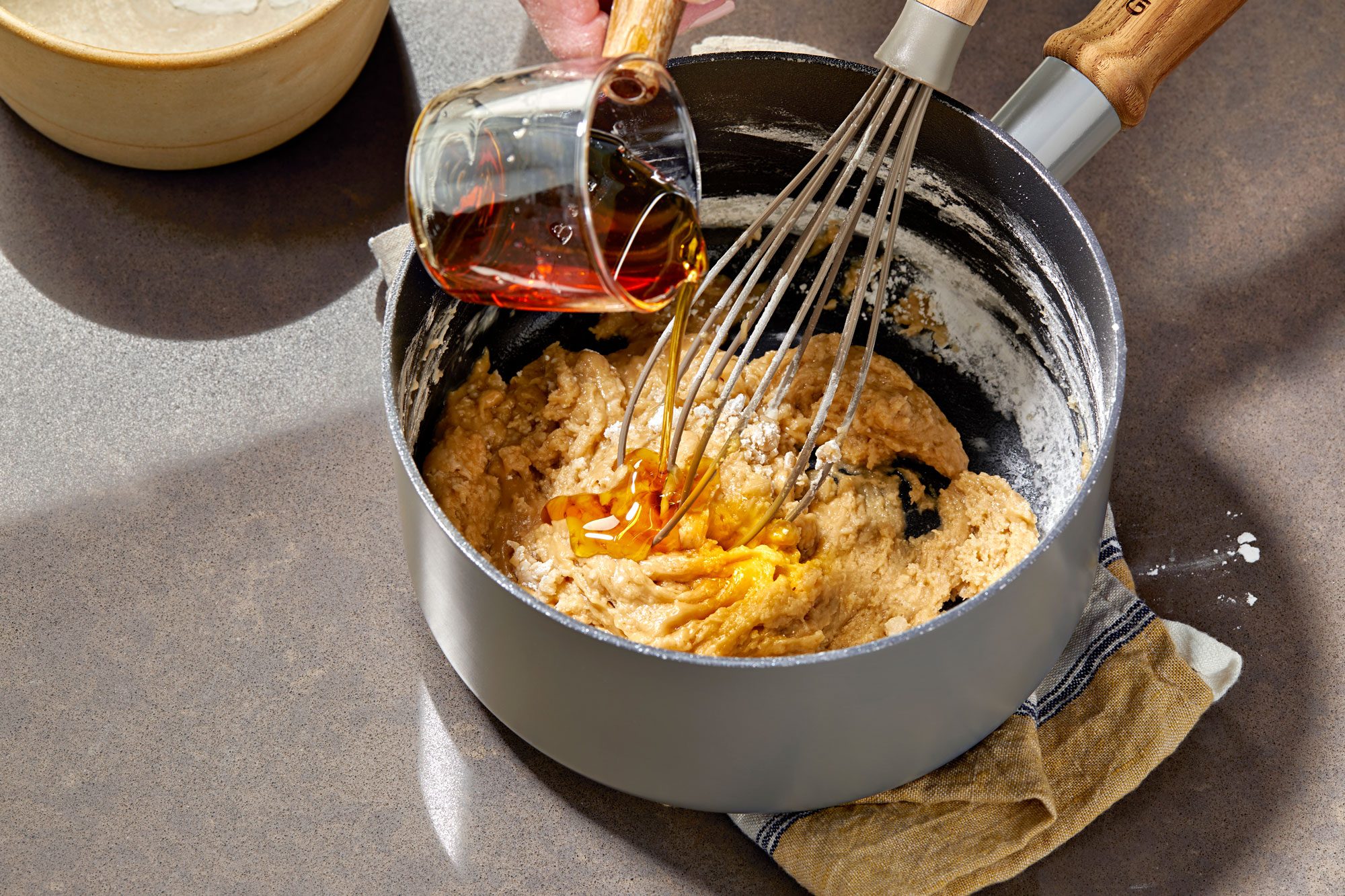 3/4th shot of a hand holds a whisk over a pot filled with brown liquid and white powder; the pot is on a beige surface, partially covered by a beige and white striped towel; a bowl of white powder and a glass of red liquid sit to the left of the pot