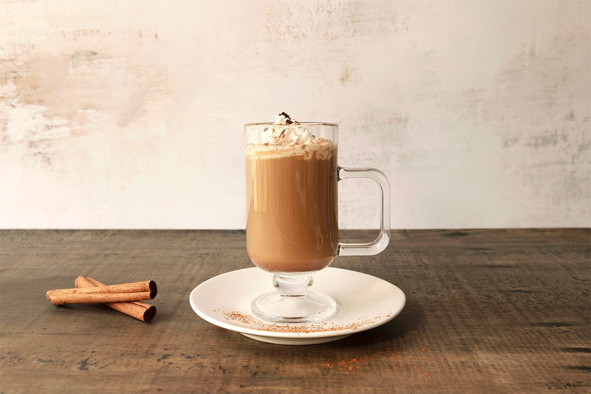 A glass mug of coffee topped with whipped cream and chocolate shavings sits on a white plate with a wooden table. Two cinnamon sticks and a small orange peel are placed on the plate beside the mug.