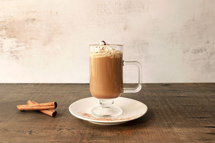 A glass mug of coffee topped with whipped cream and chocolate shavings sits on a white plate with a wooden table. Two cinnamon sticks and a small orange peel are placed on the plate beside the mug.