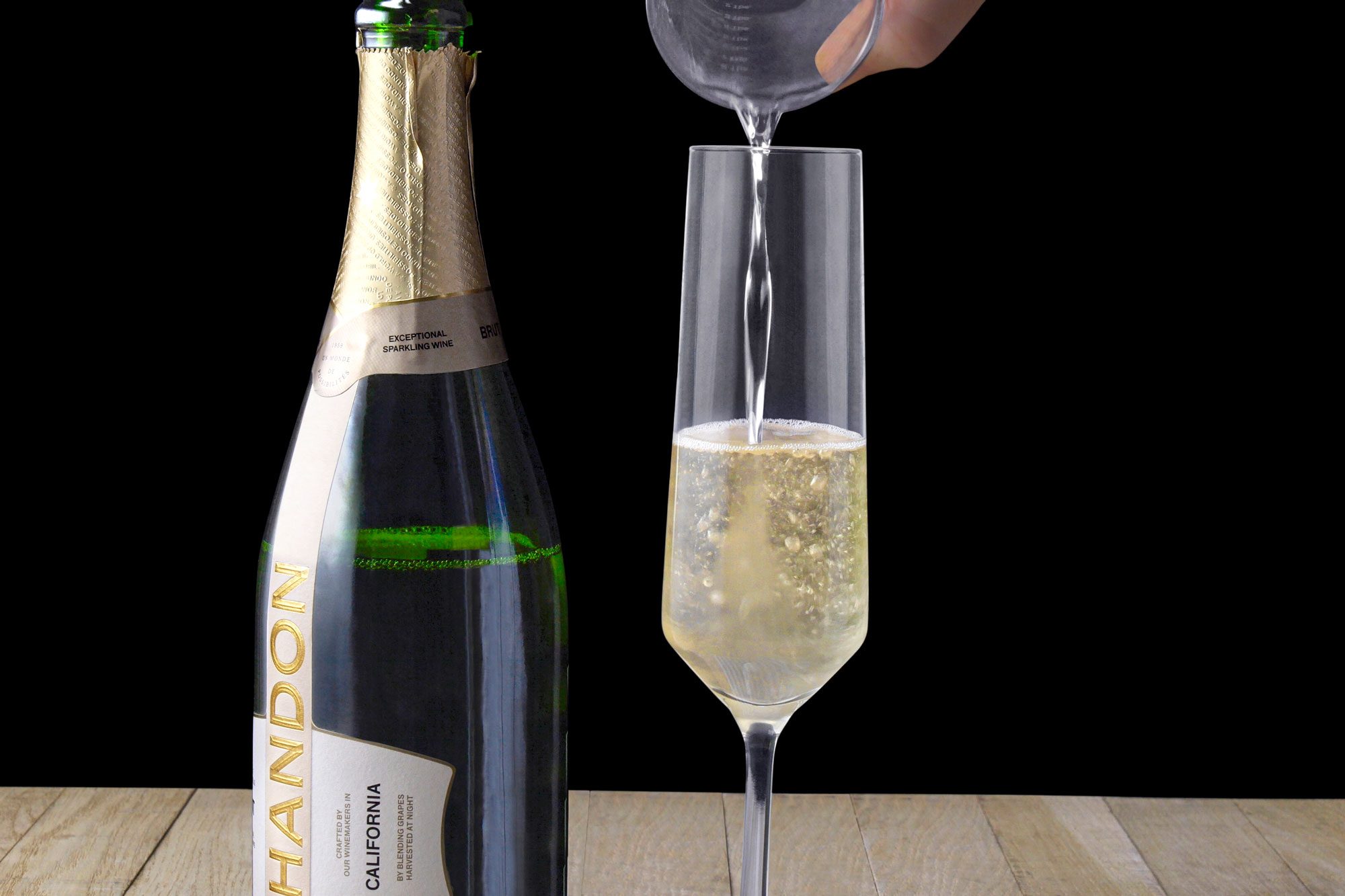 horizontal shot of a hand pouring sparkling wine from a bottle into a glass, with the backdrop being a dark