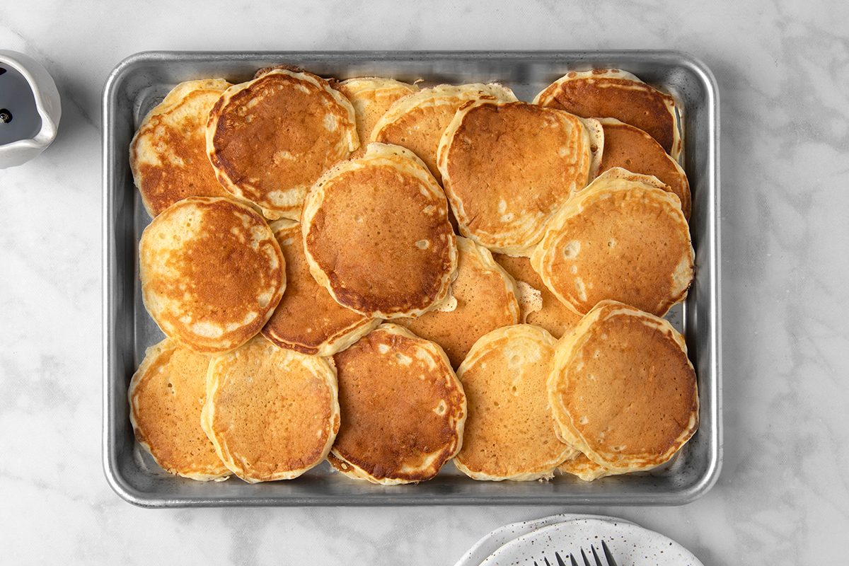A tray filled with a stack of golden-brown pancakes is placed on a marble surface. A partially visible plate with a fork lies beside the tray.