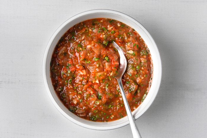 A bowl of chunky salsa sits on a light surface, filled with a vibrant mix of tomatoes, onions, cilantro, and peppers. A spoon rests inside the bowl, showcasing the salsa's fresh and colorful ingredients.
