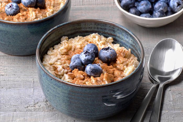 closeup shot of two bowls of oatmeal; one with a spoon and one with a bowl of blueberries, sitting on a wooden table; The oatmeal is topped with blueberries and cinnamon; The bowls are blue and the spoons are silver