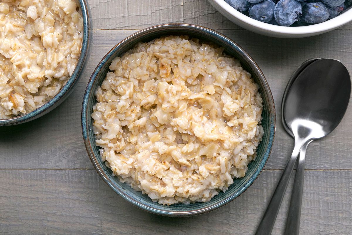 closeup shot of two bowls of oatmeal; one with a spoon and one with a bowl of blueberries, sitting on a wooden table; The oatmeal is topped with blueberries and cinnamon; The bowls are blue and the spoons are silver