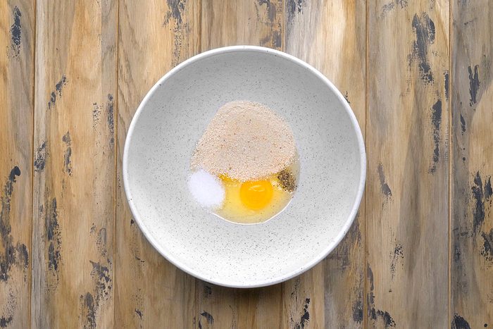 A white bowl on a wooden surface contains a raw egg, a mound of flour, a small pile of sugar, and a dash of salt, ready to be mixed.