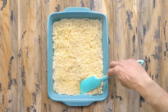 A person is using a teal spatula to spread a layer of grated potato over a dish in a blue baking pan. The pan is placed on a wooden surface with a rustic appearance.