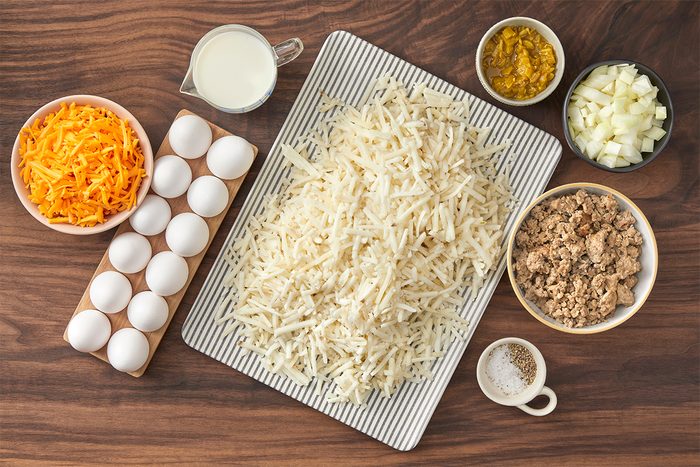 Top view of ingredients on a wooden table, including shredded potatoes, eggs, grated cheese, a cup of milk, chopped onions, ground meat, diced jalapeños, and a small bowl of salt.