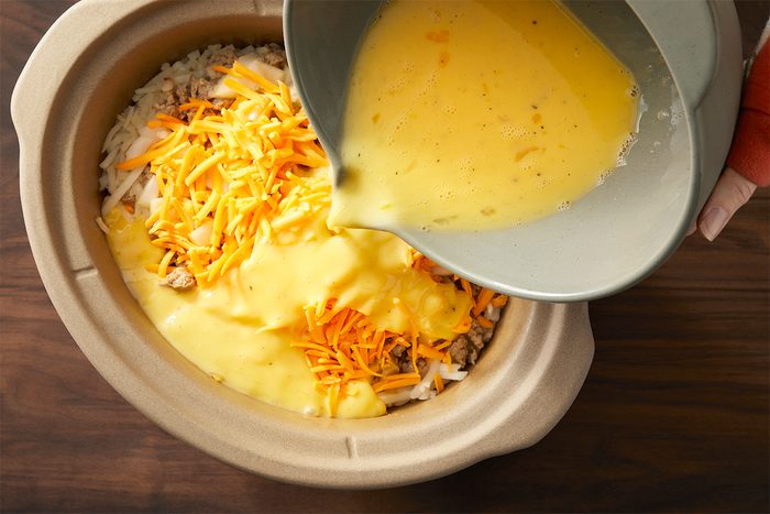 A slow cooker filled with shredded potatoes and cheese, with creamy sauce being poured in from a gray bowl. The mixture is being prepared on a wooden surface.