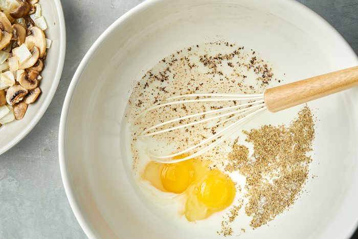 A mixing bowl contains two cracked eggs, ground spices or herbs, and a whisk. Partially visible on the side is a plate with sliced mushrooms and onions. The bowl is set on a gray surface.