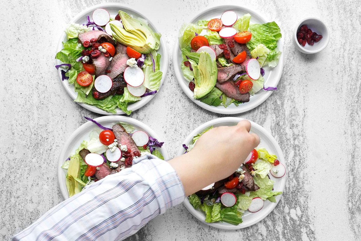 A person's hand is arranging ingredients on four plates of salad. Each plate contains lettuce, sliced radishes, cherry tomatoes, avocado, and pieces of beef. A small bowl of cranberries is placed beside the plates on a gray surface.