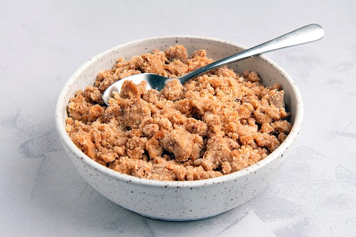 A speckled white bowl filled with crumbly brown sugar sits on a light gray marble surface. A silver spoon is resting in the sugar, angled slightly to the right.