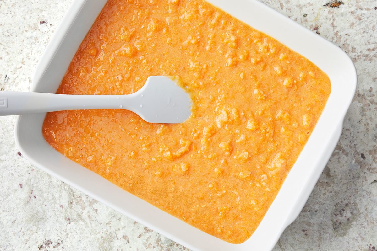 A white rectangular dish filled with an orange, lumpy mixture that resembles pumpkin batter. A gray spatula rests inside the dish, partially submerged in the batter, on a speckled countertop.