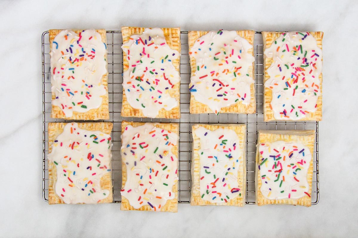 Eight homemade pop tart pastries on a wire cooling rack on top of a white countertop, with white icing and rainbow sprinkles