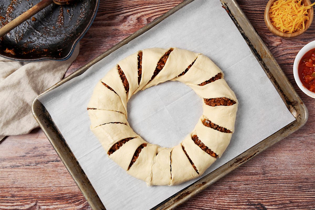 overhead shot of a baking sheet lined with parchment paper and a ring of dough that is filled with a mixture of ground meat and vegetables; the dough is cut into slices and twisted; the baking sheet is on a wooden surface and there are two bowls of ingredients on the side; one bowl contains shredded cheese and the other contains a tomato salsa;
