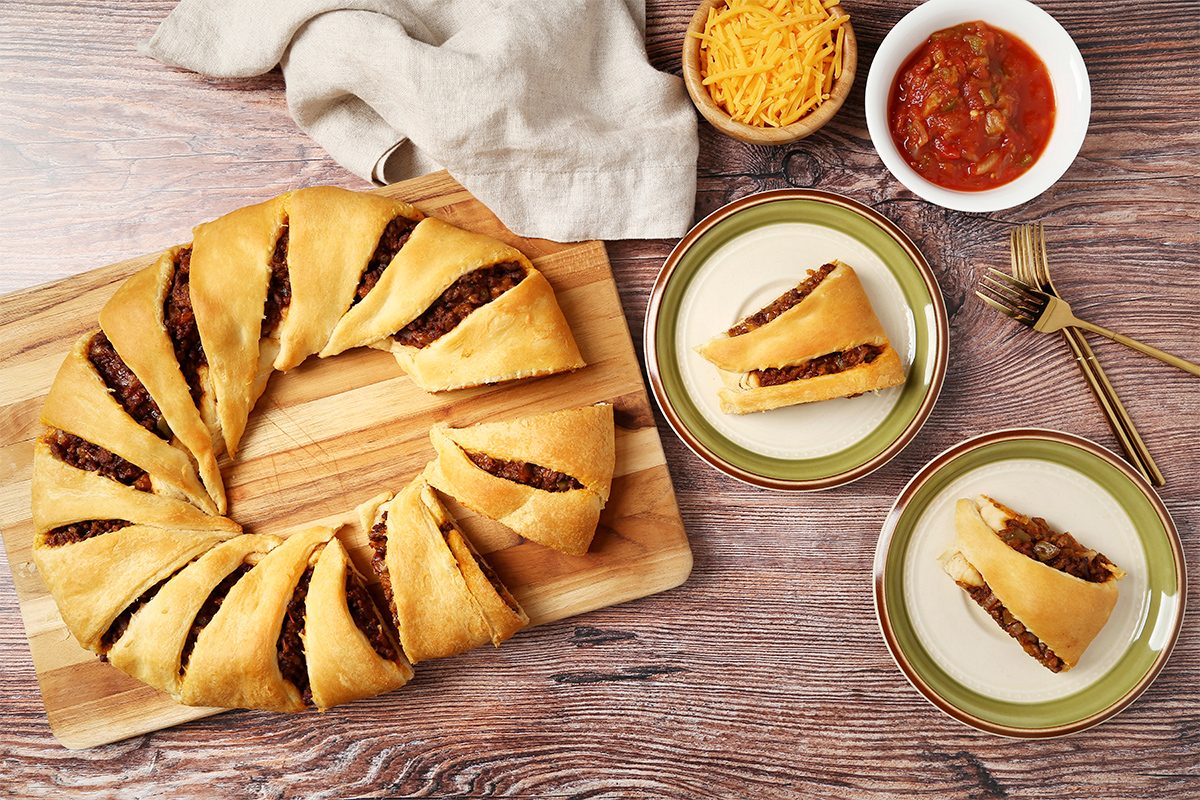overhead shot of a wooden cutting board is in the center with a golden brown pastry ring with filling inside, a portion of it is separated into 8 triangular pieces; a white ceramic bowl with red salsa, a bowl with grated cheese, and two green and brown patterned plates, each with a single piece of the pastry ring; golden forks are lying on the side; the background is a wooden surface with a light brown linen cloth to the left