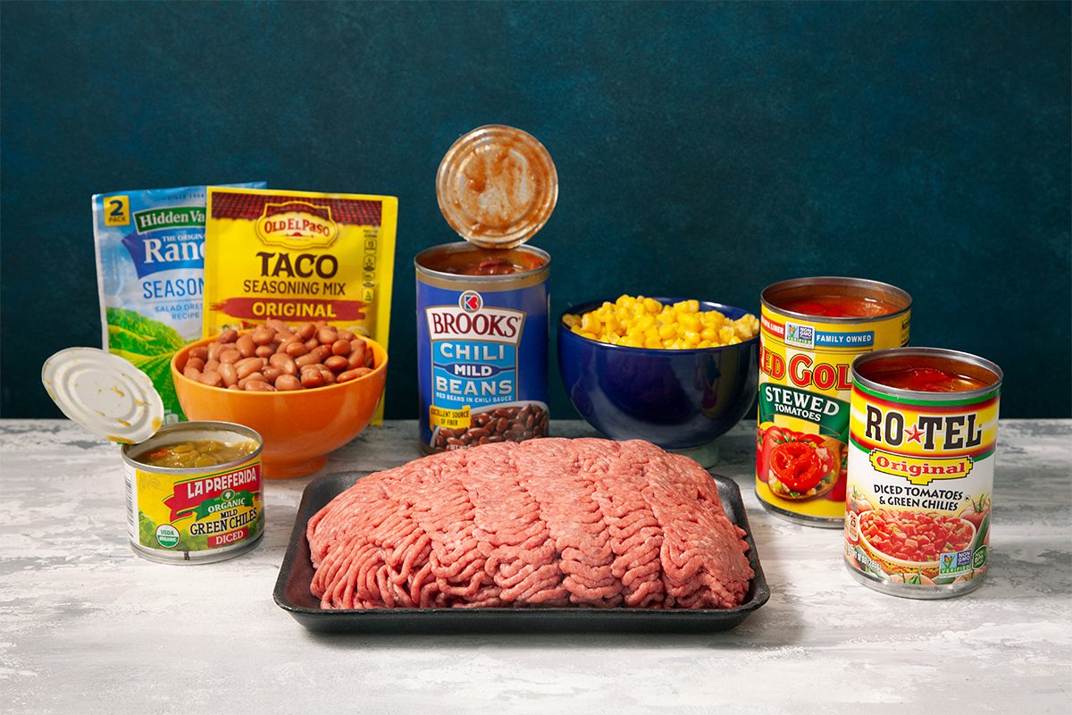 A selection of food items on a table: ground meat on a tray, cans of beans, corn, diced tomatoes, green chilies, and ranch dressing mix, plus a packet of taco seasoning. A blue bowl is filled with corn.