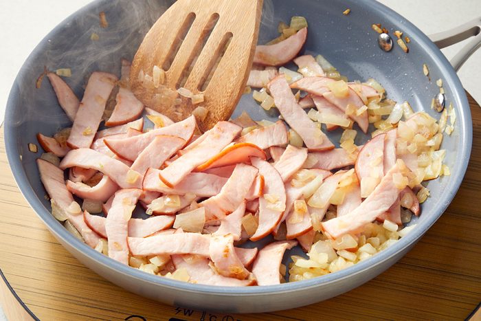 Canadian bacon added into the cooked onions in a large skillet with a ladle to stir