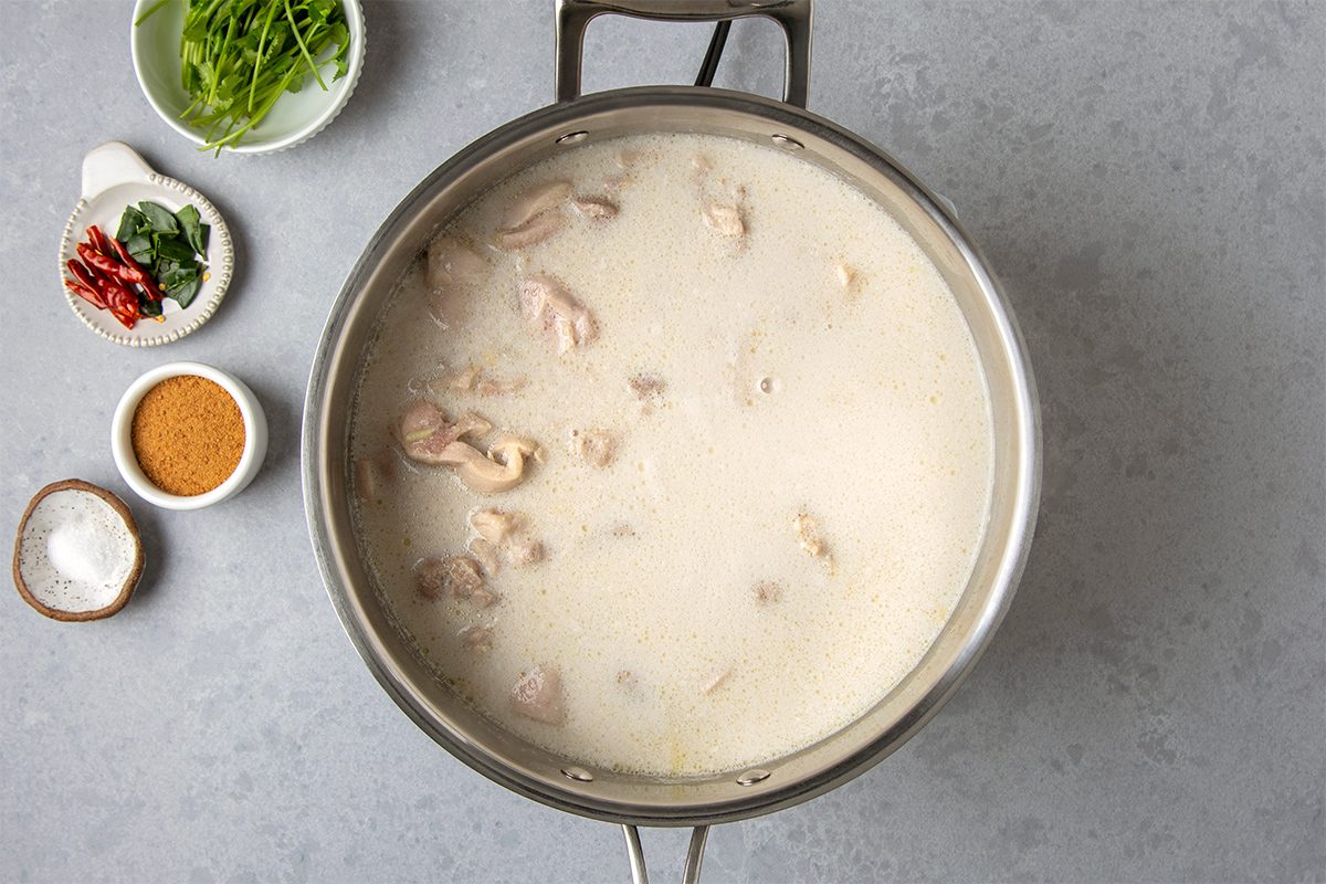 A pot of creamy soup with chunks of meat, placed on a gray countertop. To the side, there are small bowls containing spices, herbs, and sliced red chilies.