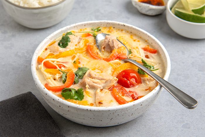 A bowl of creamy soup with sliced red peppers, chicken chunks, and herbs, garnished with a lime. A spoon rests inside the bowl. Nearby are small bowls with additional garnishes and a napkin on a gray surface.