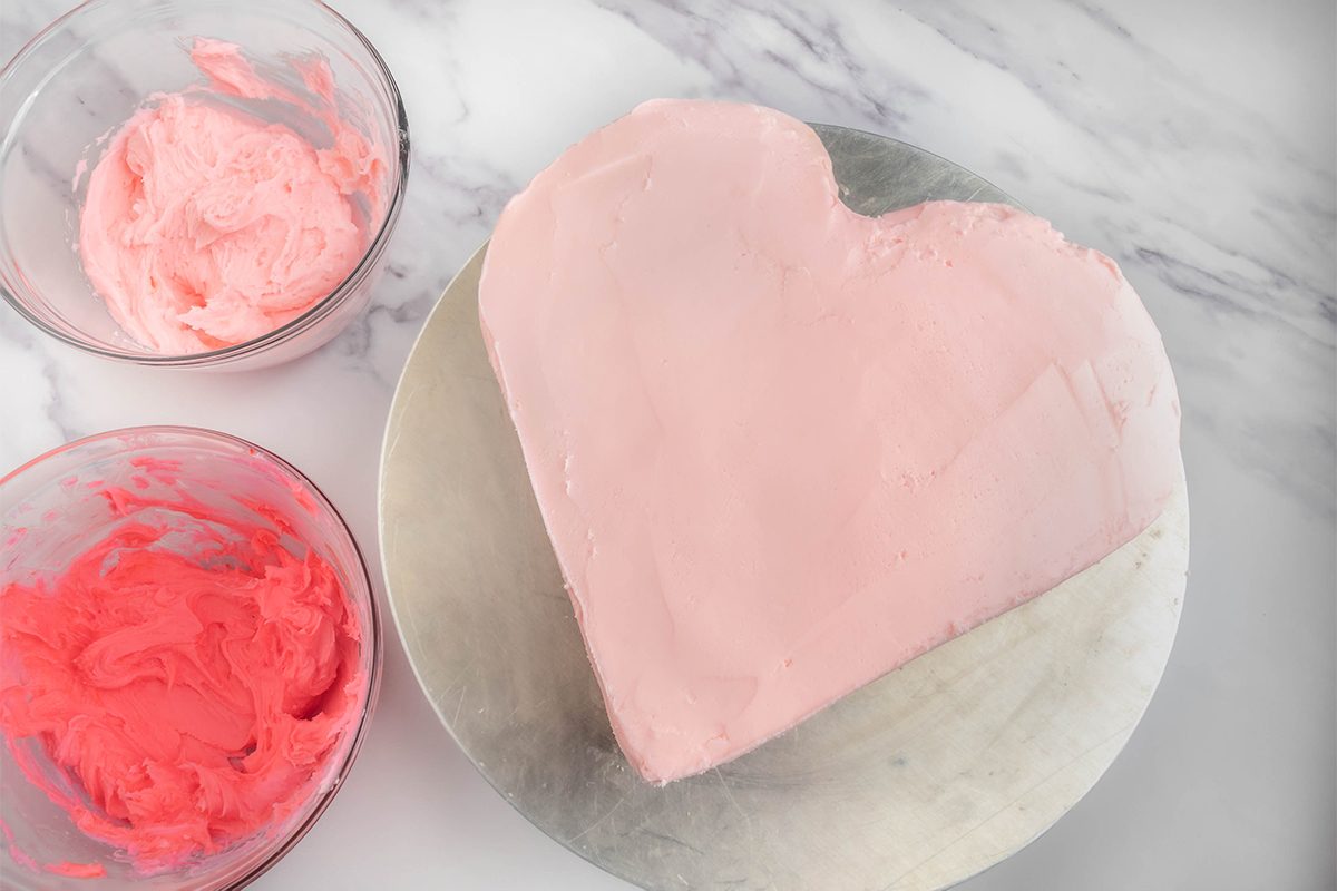 A heart-shaped cake with light pink frosting is placed on a round board. Two bowls with pink frosting, one darker and one lighter, are beside the cake on a marble surface.