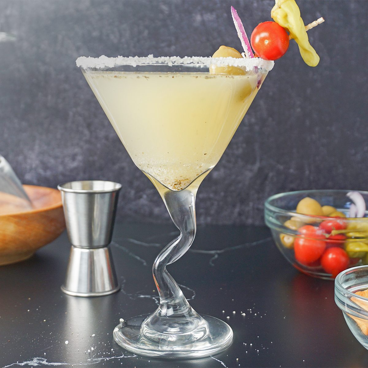 A martini glass with a salted rim holds a light yellow cocktail, garnished with a cherry, olive, onion, and pepper. The glass has a twisted stem. In the background, there are ingredients and a measuring jigger on a dark surface.