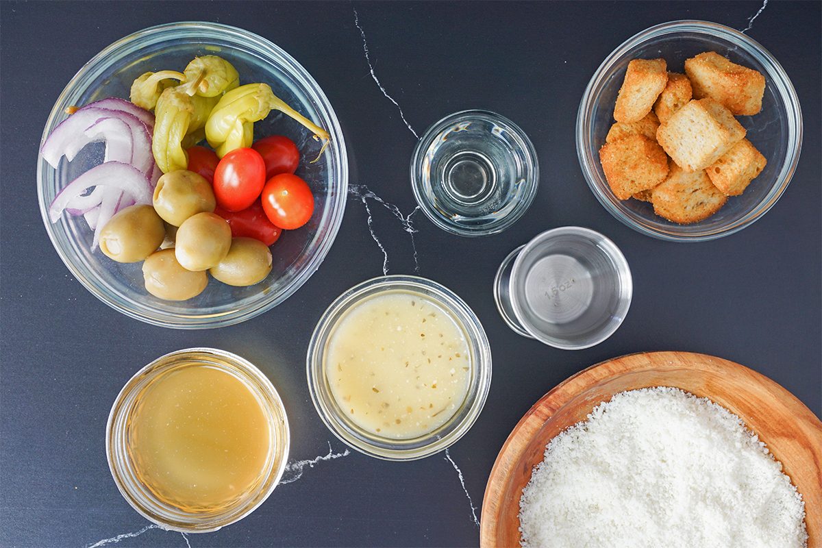 Bowls on a dark surface containing sliced onions, cherry tomatoes, olives, green chili peppers, croutons, a liquid sauce, clear liquids, a cheese-like substance, and a wooden plate with grated cheese.