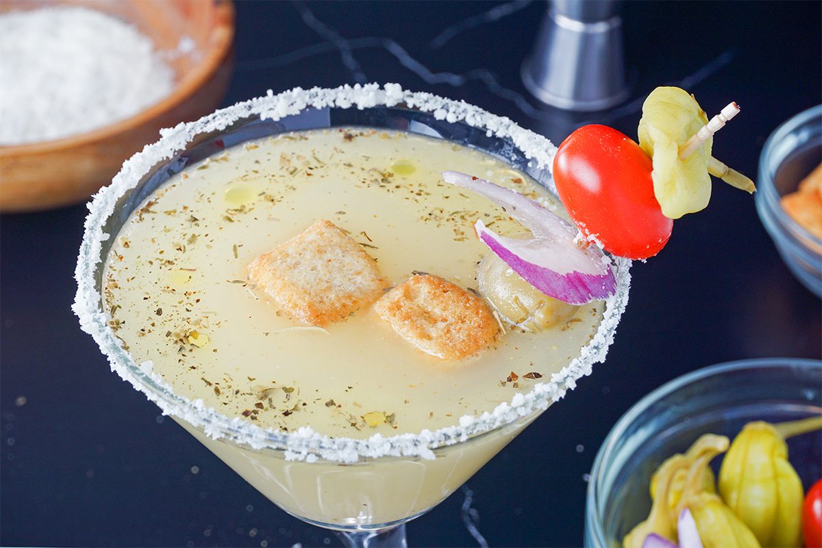 A cocktail glass with a salted rim contains a savory drink garnished with croutons and herbs. A skewer with a cherry tomato, pickle, and pepperoncini rests on the glass. Surrounding the glass are bowls with ingredients on a dark surface.