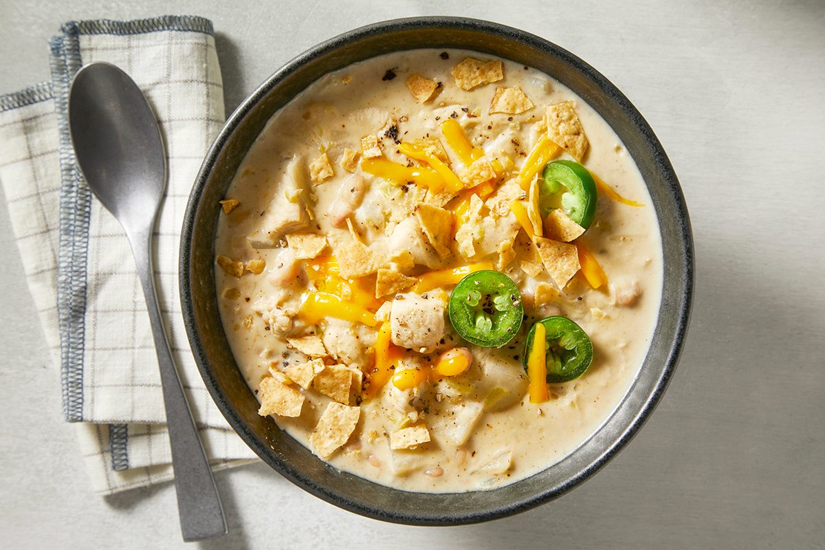 A bowl of creamy soup topped with shredded cheese, sliced jalapeños, and crushed tortilla chips. A spoon rests on a checkered cloth beside the bowl, on a light-colored surface.